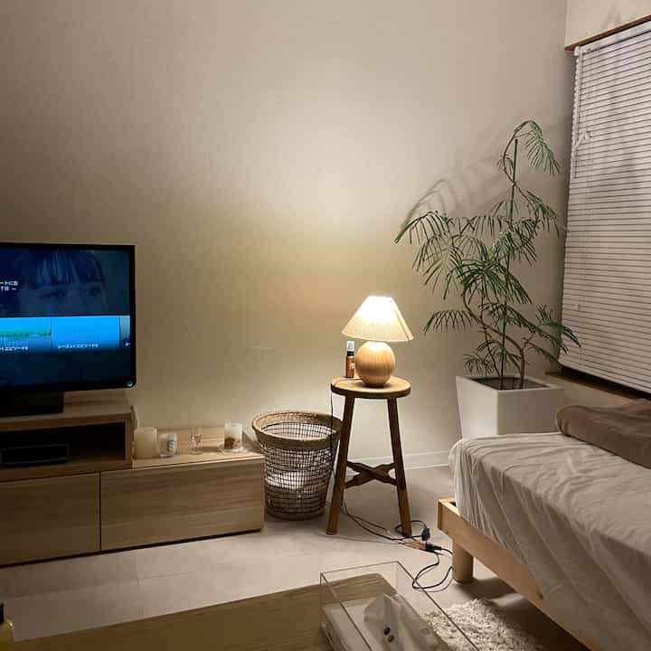 ベージュの壁と木目調家具が配置された一人暮らし用の寝室、テーブルランプと大きな観葉植物が暖かな照明の下で落ち着いた雰囲気を演出
