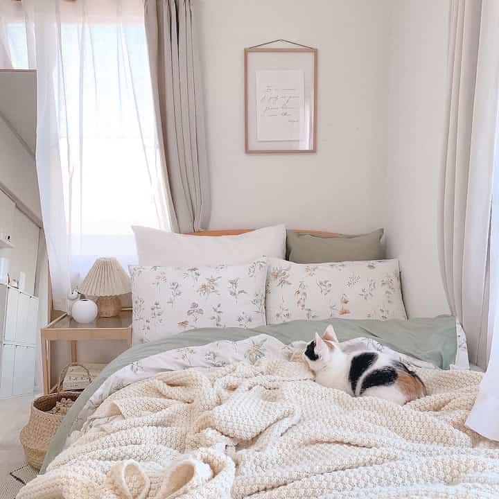 White and beige toned bedroom featuring floral pillows and a knitted blanket with a cat resting, creating a cozy atmosphere
