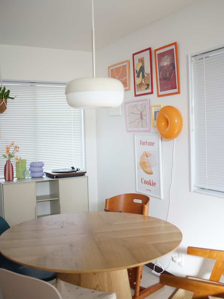 白い壁のダイニングルーム、木製の丸いテーブル、カラフルなポスターとオレンジ色の壁照明が配置されたシンプルなインテリア空間