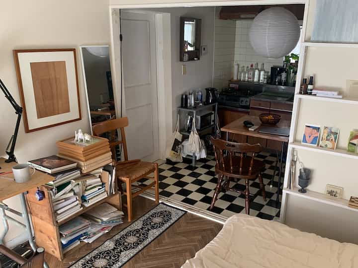 White and brown toned studio apartment featuring kitchen and living area with wooden furniture and checkered flooring, giving a cozy atmosphere
