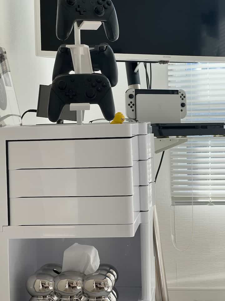 白を基調としたリビング、ゲームコントローラー収納ラックとテレビ、ブラインドが配置された清潔な空間