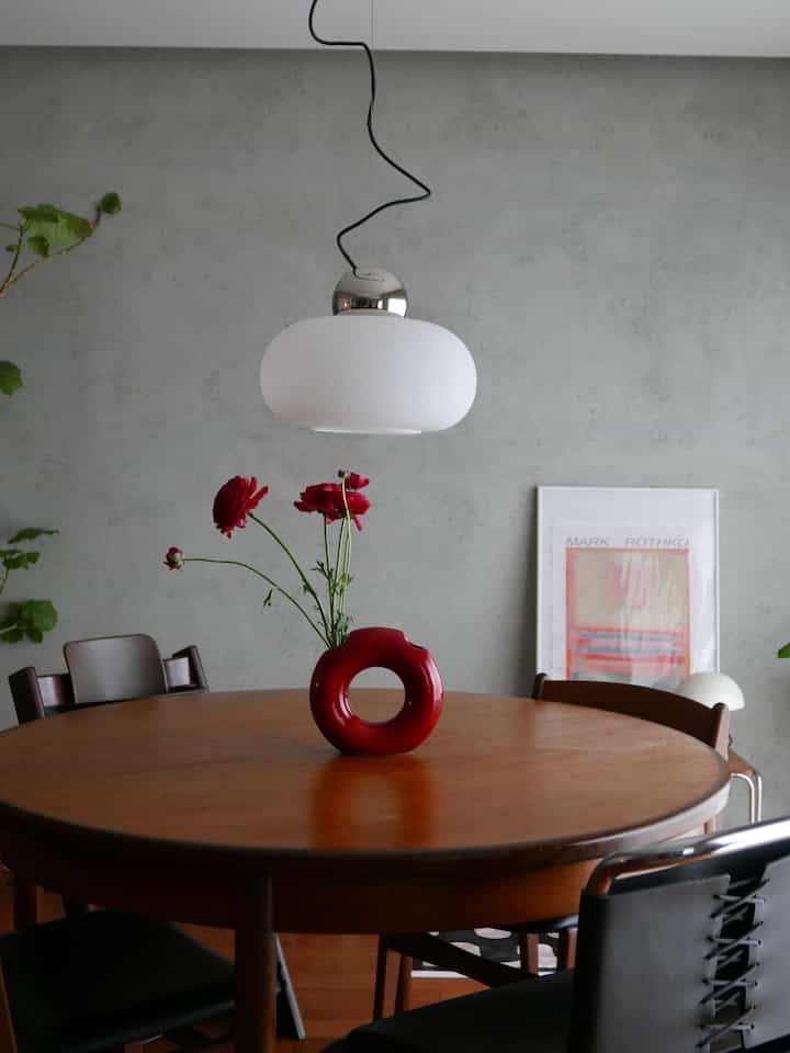 グレーのコンクリート壁と木製の丸いダイニングテーブル、赤い花瓶が置かれたモダンなダイニングルーム