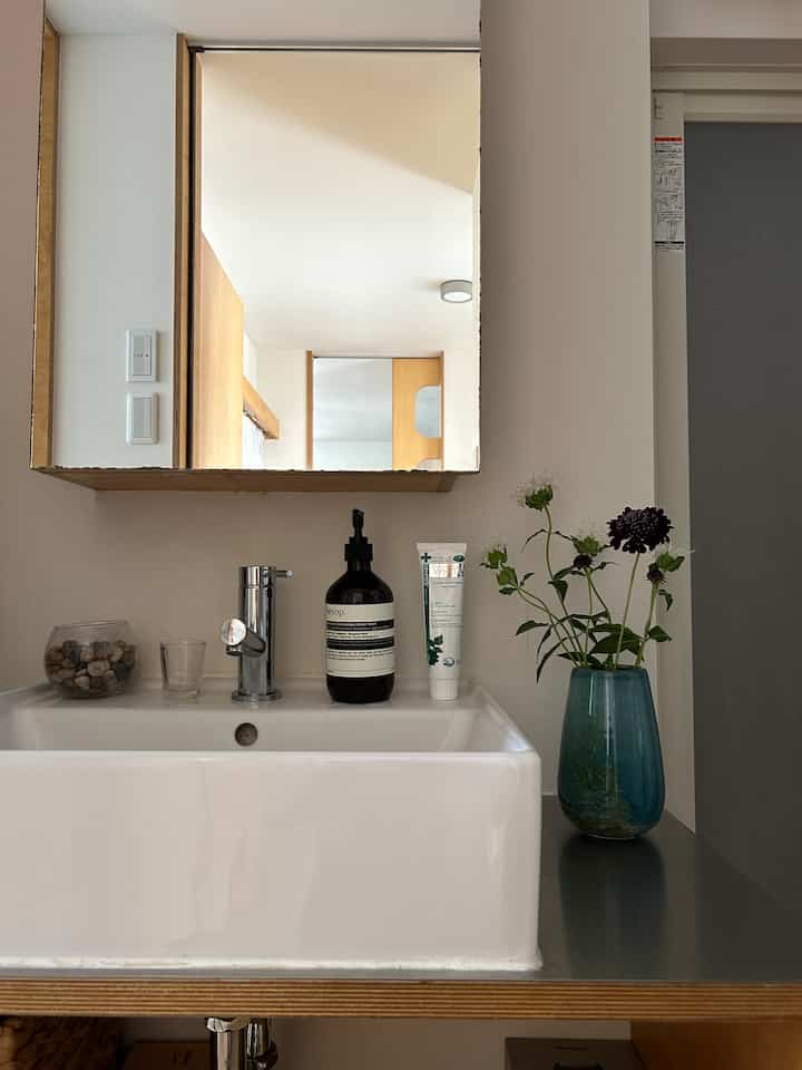 白とナチュラルカラーが調和したお手洗い、シンプルな洗面台と木製枠の鏡、花瓶に生けられた花が配された落ち着いたインテリア