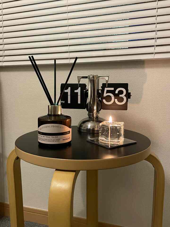 透明な氷型キャンドルとダークブラウンのディフューザーを置いたモダンなサイドテーブルと白いブラインドが配された雰囲気のある空間