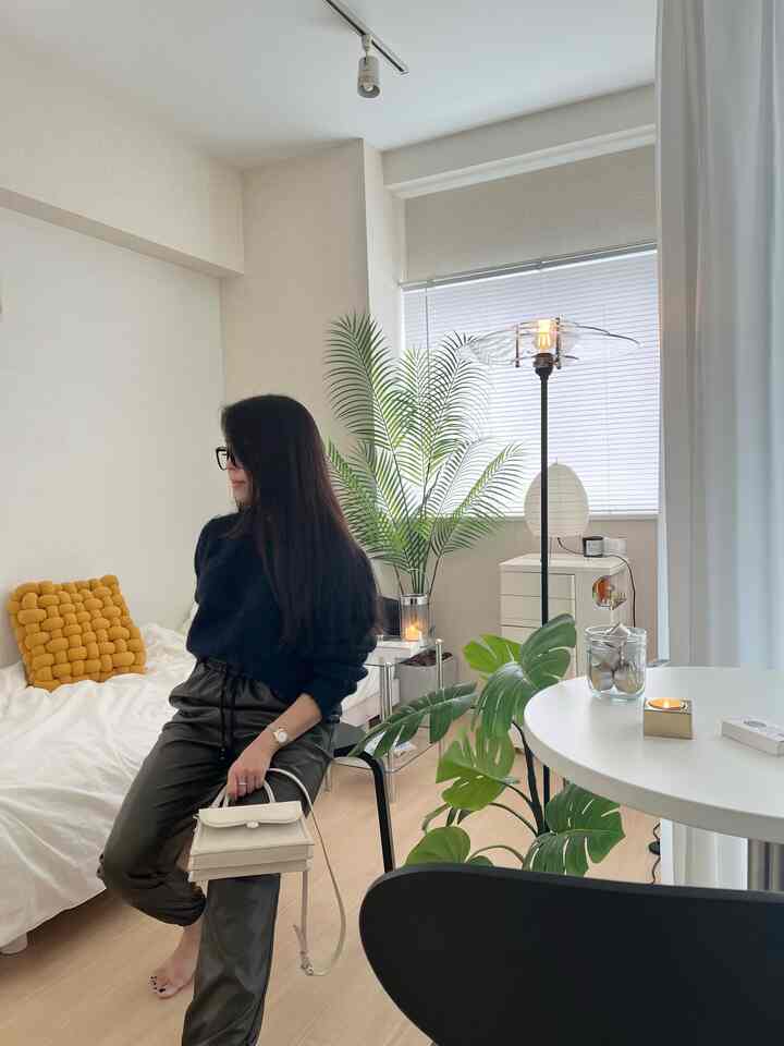 A narrow bedroom in white and brown tones featuring a white bed with a yellow cushion, plants, and a modern floor lamp