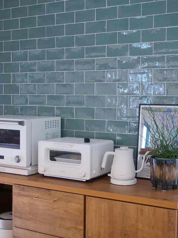 光沢のあるグリーンタイルの壁と木目調の収納が配置されたミニマルモダンなキッチン、ホワイトのBALMUDA家電が調和している