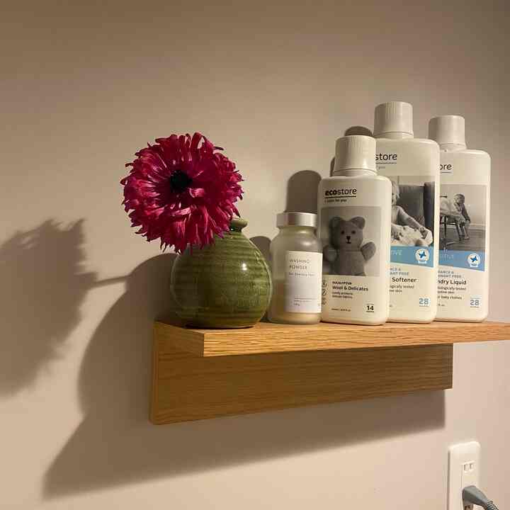 Natural wood tone shelf with red flower and bathroom detergents arranged on white wall, creating simple bathroom space