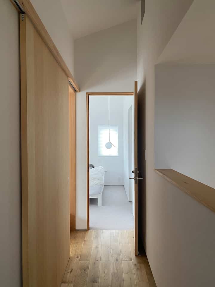 ナチュラルなブラウンの廊下と白い壁、カーペットが敷かれた寝室がつながる、シンプルで落ち着いたインテリア