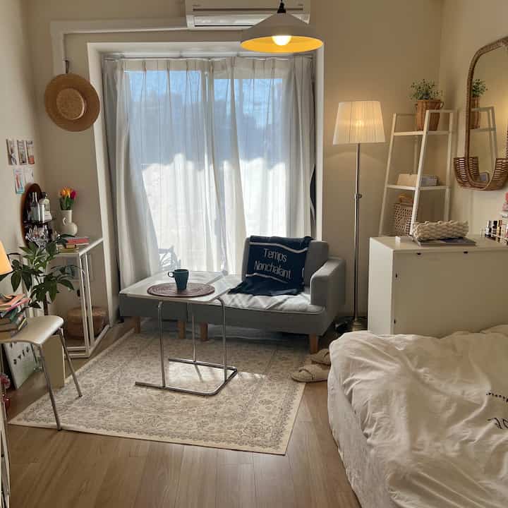 Natural wood tones and white curtains in a compact studio bedroom, featuring a gray sofa and side table creating a cozy atmosphere