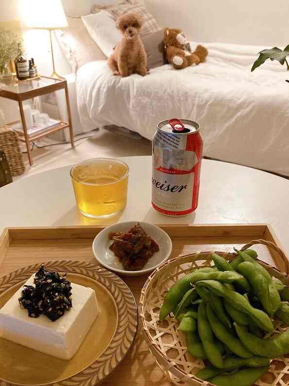 ホワイトとナチュラルカラーのリビング、ベッドの上に小さなペットの犬とテディベアが置かれ、テーブルにはビールとおつまみが並ぶ雰囲気のある空間