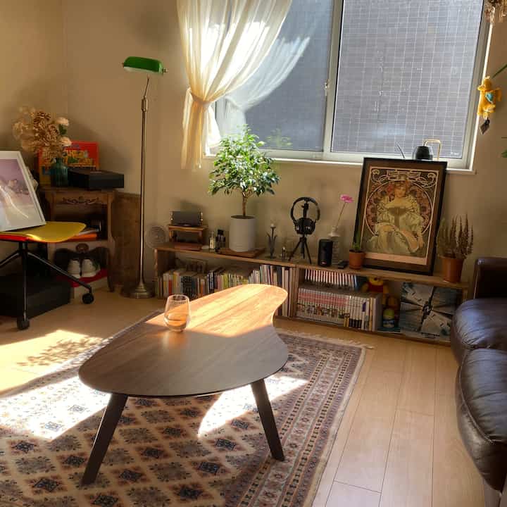 ナチュラルカラーのリビング、個性的なコーヒーテーブルとラグ、窓辺に配置された植物とポスターが調和した雰囲気のあるインテリア