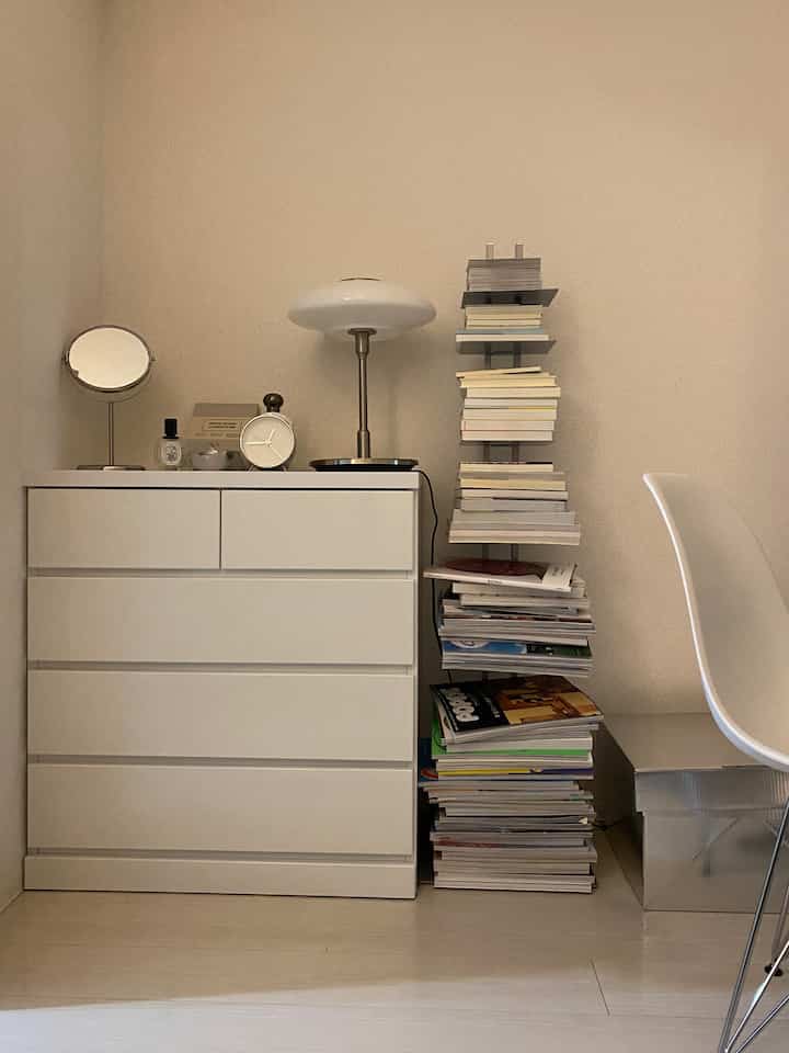 白とベージュの色調の狭い部屋で、白いチェストと本棚、テーブルランプが配置されたミニマルな空間