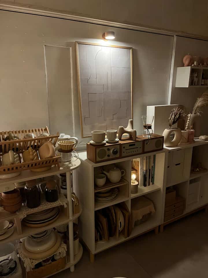 ベージュとホワイトトーンのナチュラルスタイルのホームカフェ空間、Atelier Cphのポスターと間接照明が際立つ収納棚とオブジェを配置したインテリア