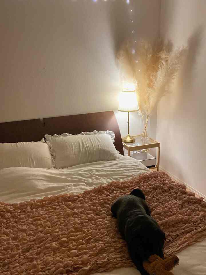 暖かいナチュラル調の寝室、中央にベッドとピンクのベッドカバー、右側に灯りのついたテーブルランプと花瓶が配置された印象的な空間