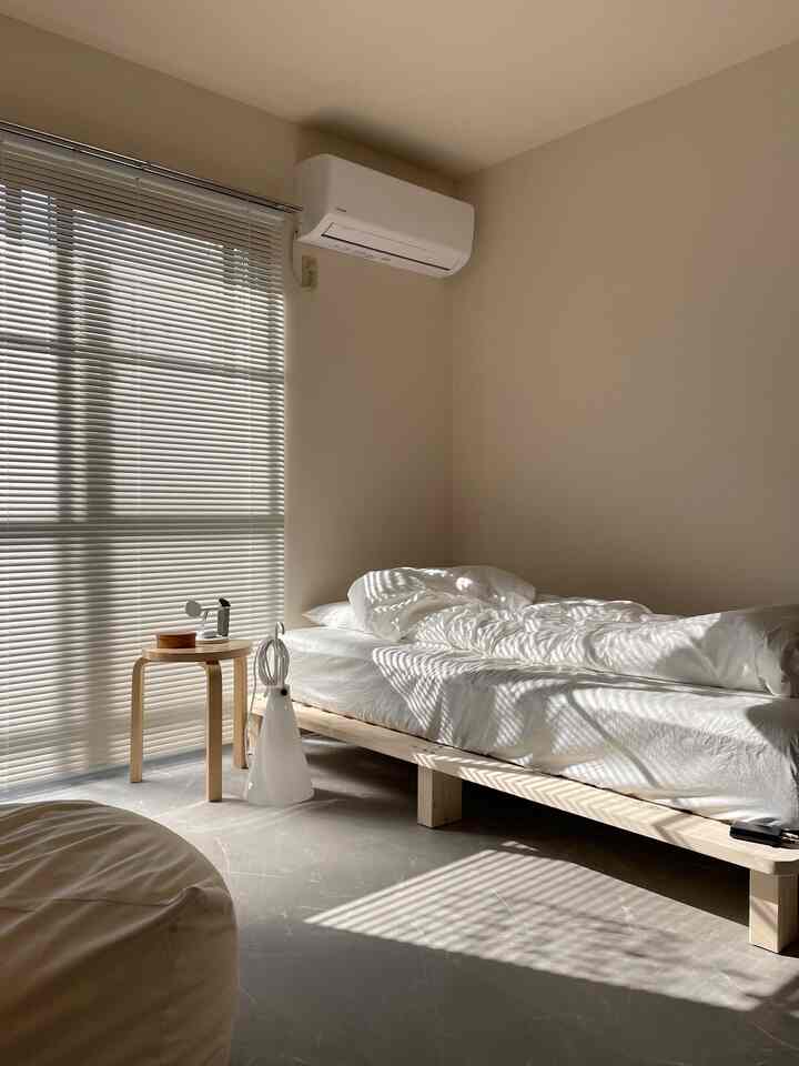 白とベージュのトーンが映える8畳の1K部屋、シンプルな木製ベッドフレームとスツールが配されたモダンでミニマルな空間