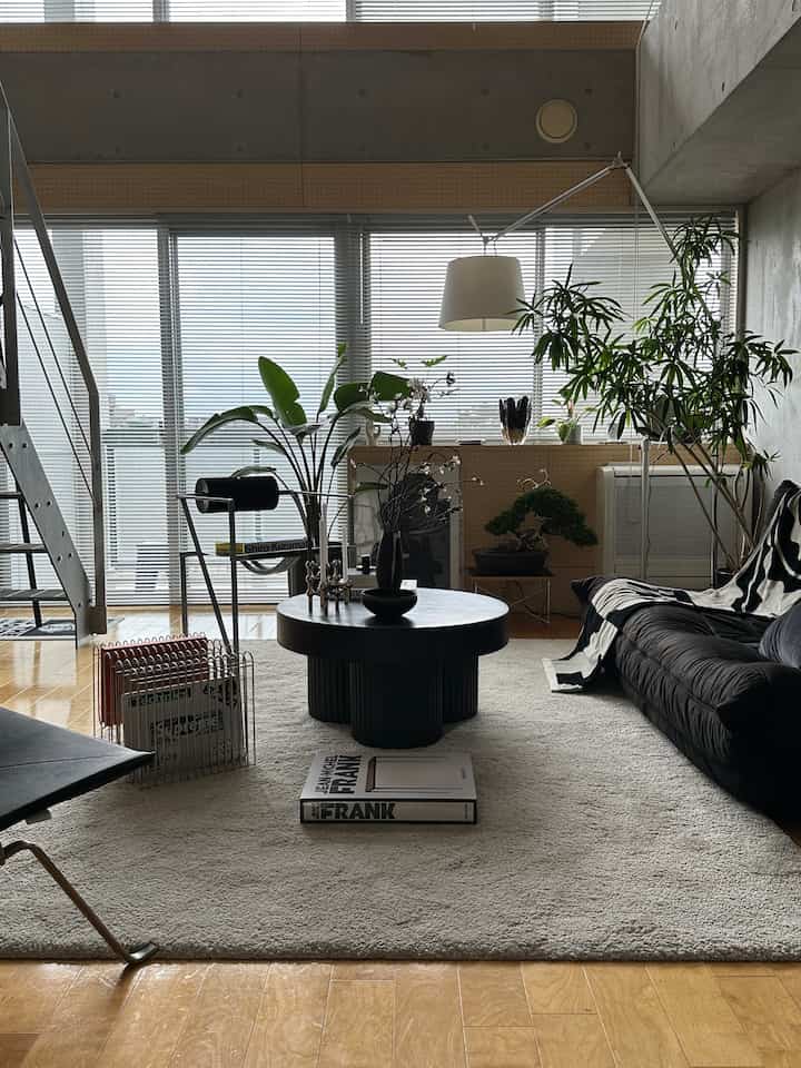 Natural tone living room featuring a black circular coffee table on an ivory rug and a black sofa with plants on the right, creating a modern ambiance