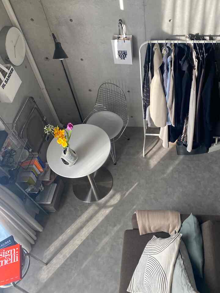 白とグレーのトーンのワンルームリビング、丸テーブルとワイヤーチェア、コートハンガーが配置されたモダンで明るい空間