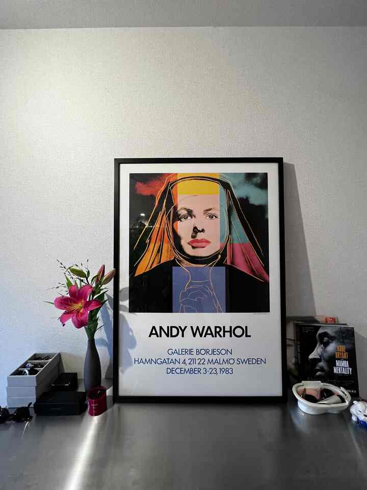 A minimalist desk space with a white wall background featuring a central Andy Warhol poster and a vase with flowers on the left