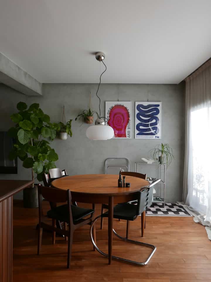 グレーのコンクリート壁と木目調床のミッドセンチュリーモダンなダイニング、丸いダイニングテーブルと椅子、ペンダントライト、植物が配置された自然な雰囲気のあるインテリア