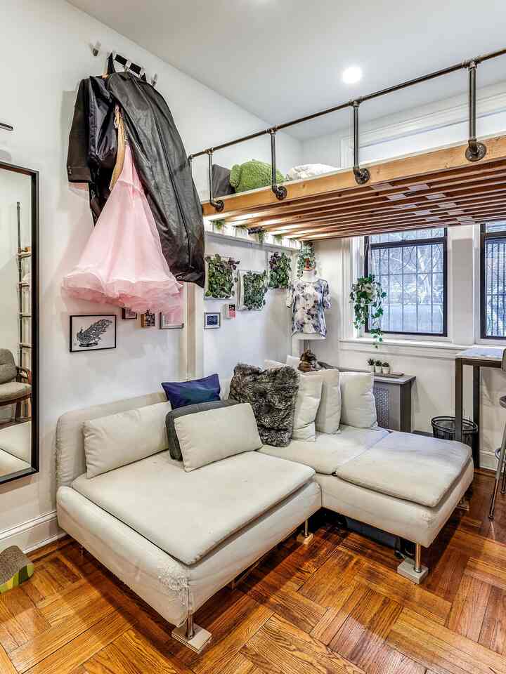 A white and wood-tone studio apartment loft living room featuring a white sectional sofa, wall-mounted plants, and a wooden loft bed for a cozy atmosphere