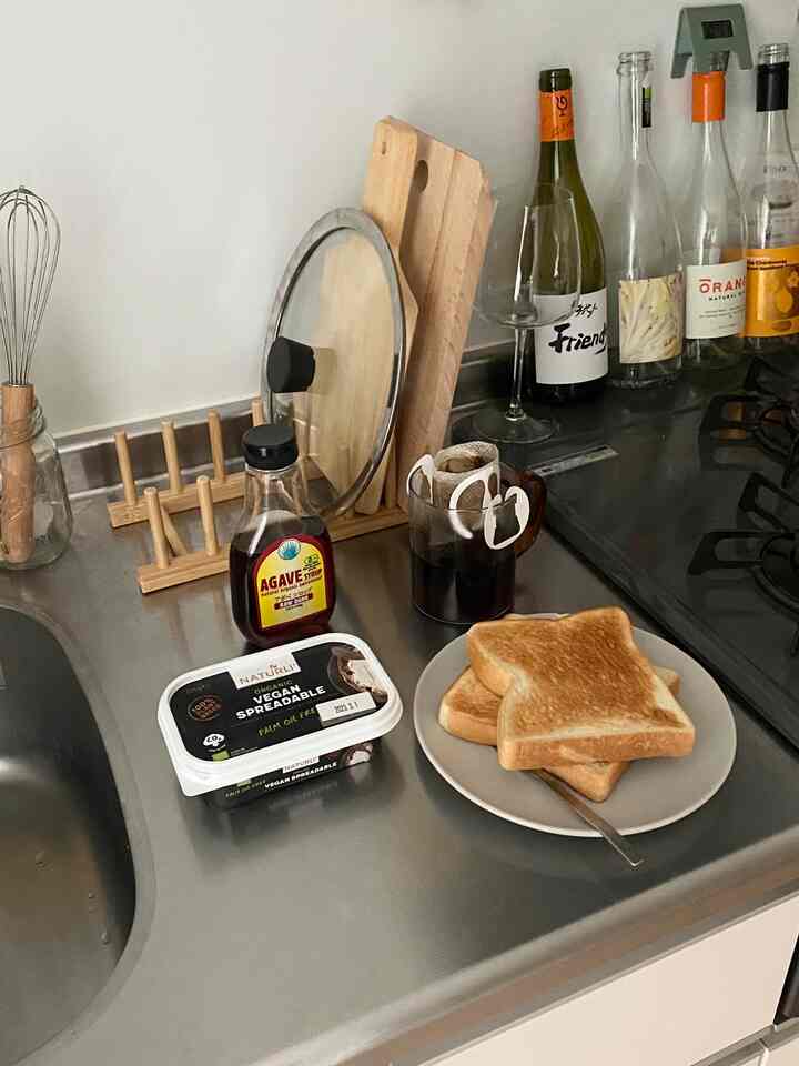 ナチュラルな色調のキッチン、ステンレス製カウンターにトースト、コーヒー、皿、木製まな板が配置された簡単な朝食の雰囲気のあるインテリア