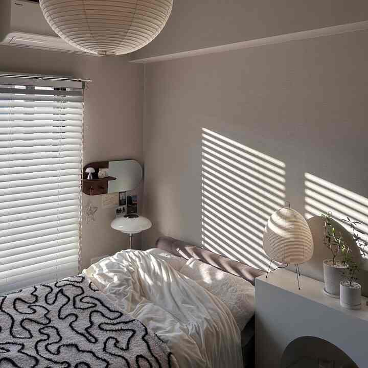 A soft gray and white toned bedroom featuring blinds-covered window, cozy bed, and warm winter sunlight casting shadows