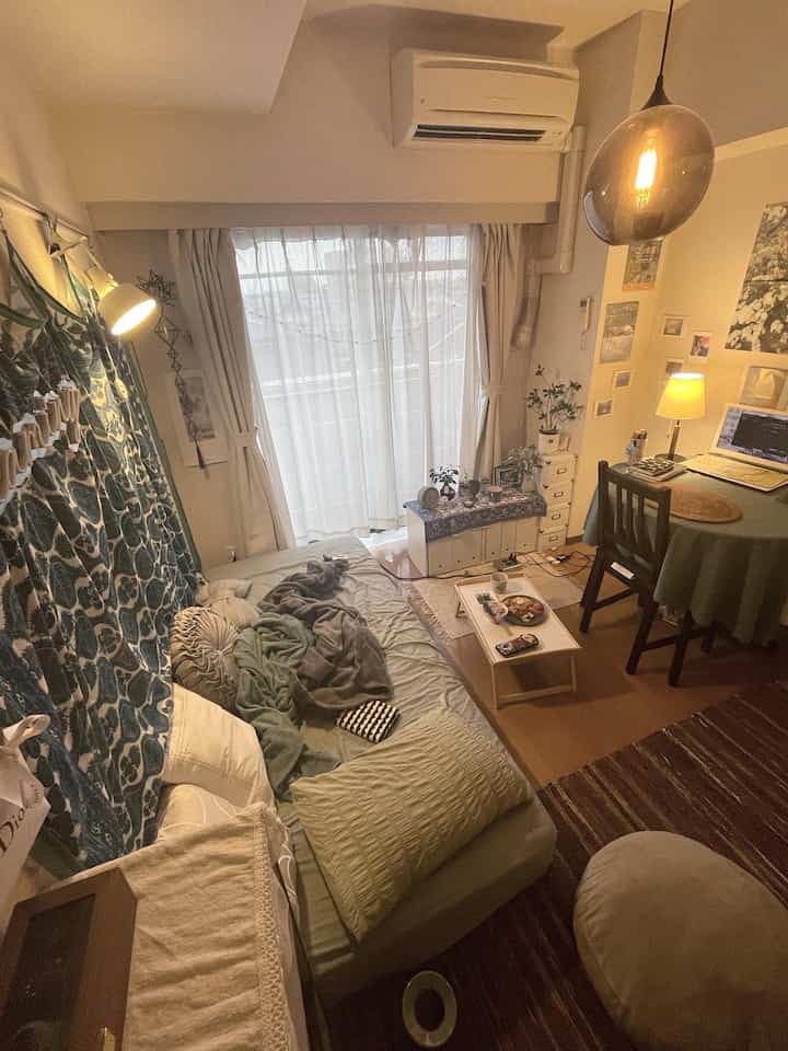 Beige-toned studio apartment bedroom featuring a sofa bed, plants, small coffee table, and a workspace with warm cozy atmosphere