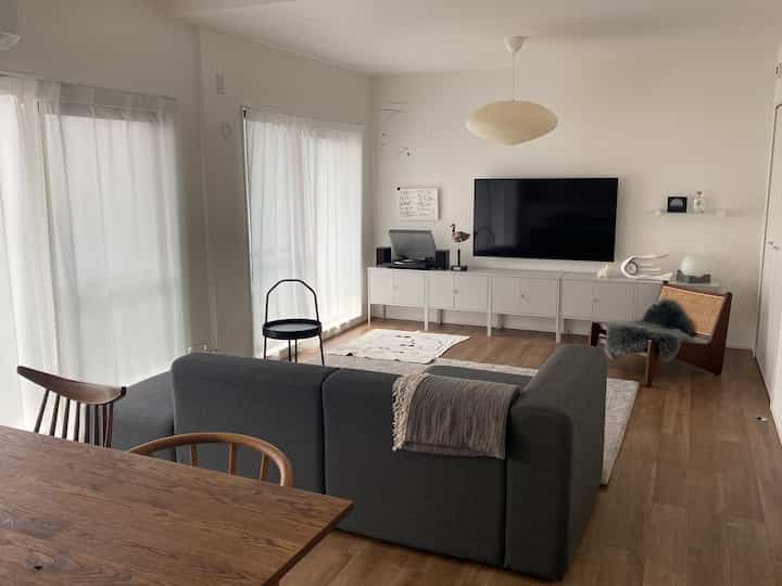 ライトブラウンの木製床と白い壁のリビング、中央にグレーのソファとケーンのアームチェアが配置されたモダンでナチュラルな印象的な空間