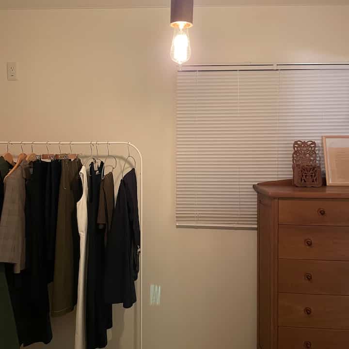 Dressing room with white walls and a brown wooden chest of drawers, featuring a pendant light and white blinds for a neat atmosphere