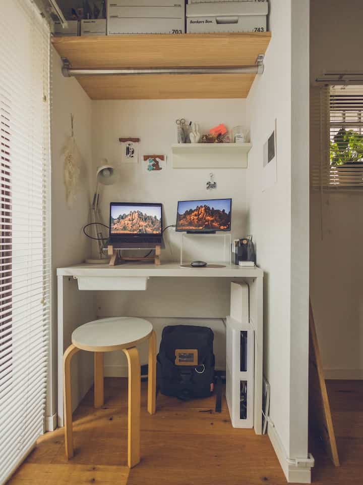 A white-walled, wood-toned small home office featuring a desk with a laptop and monitor neatly arranged