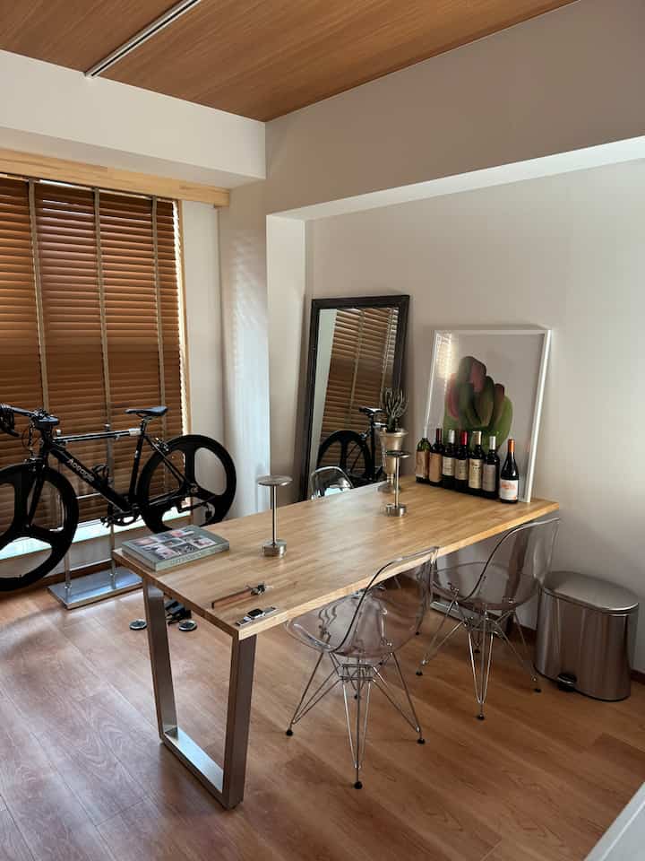 Natural warm-toned single household dining room featuring a wooden table with transparent chairs, large wall mirror, and a bicycle by the window