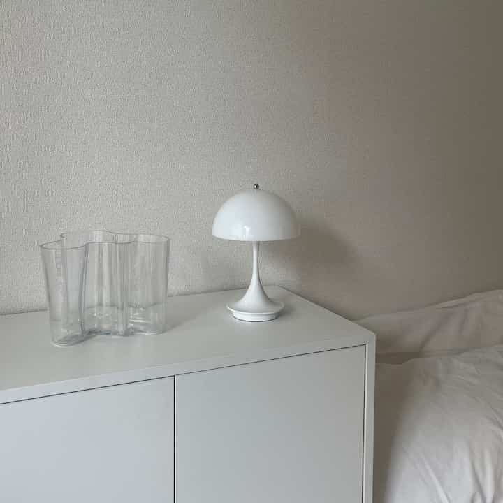 清潔感のある白い収納キャビネットの上に、白いテーブルランプと透明な花瓶が置かれたシンプルなワンルームのインテリア