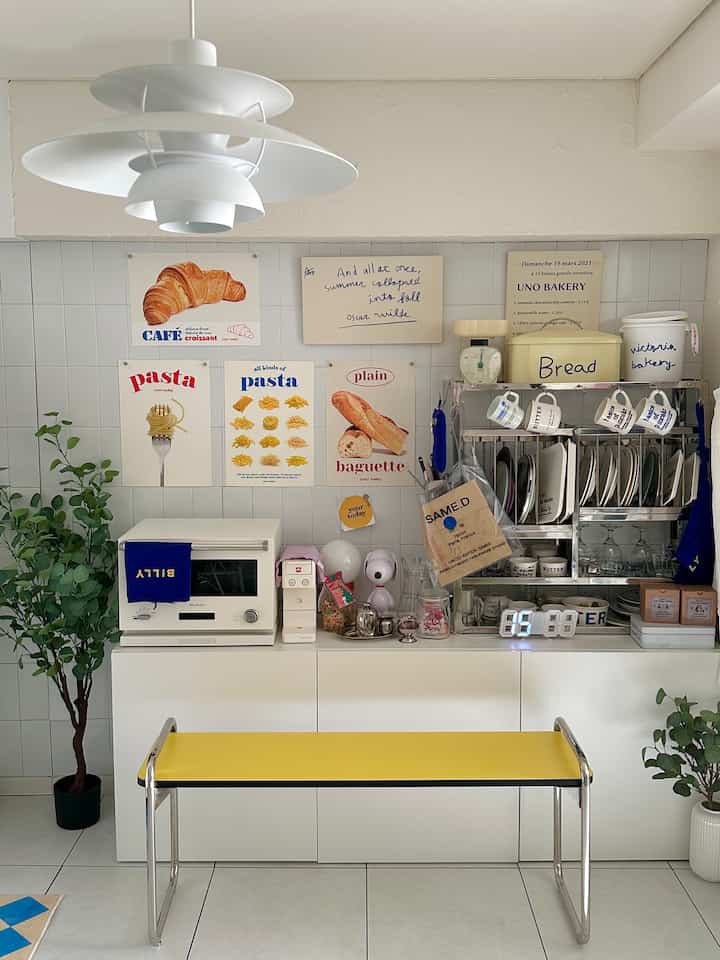 明るい白と黄色のトーンのキッチン空間、壁に多様なポスターが並び、棚にはキッチン用品が配置された清潔感のある雰囲気のホームカフェ風インテリア