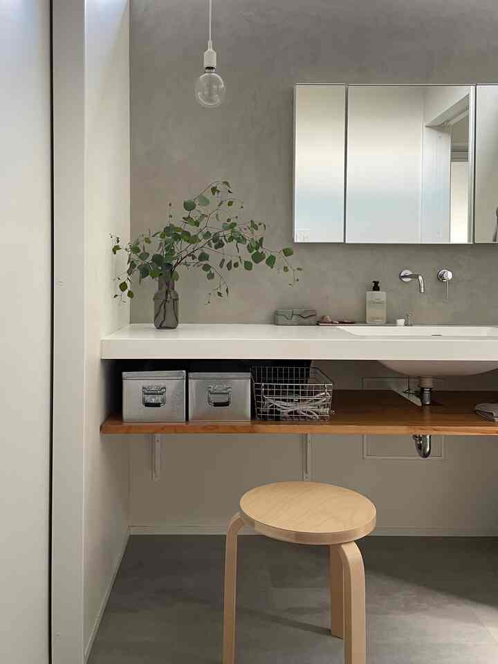 グレーの壁と白い洗面台が配置された浴室、木製スツールと棚、そして緑のユーカリの枝が置かれたシンプルで自然な雰囲気のインテリア