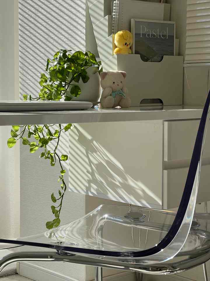 White and transparent toned workspace featuring desk, transparent chair, plant, and poster with a clean atmosphere