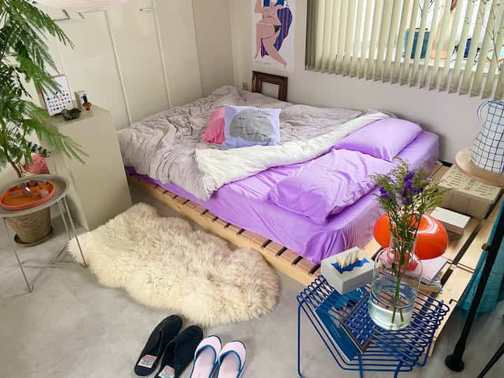 パープルとベージュのトーンの寝室、木製ベッドと猫柄クッション・掛け布団カバーが配置された自然で落ち着いた雰囲気のインテリア