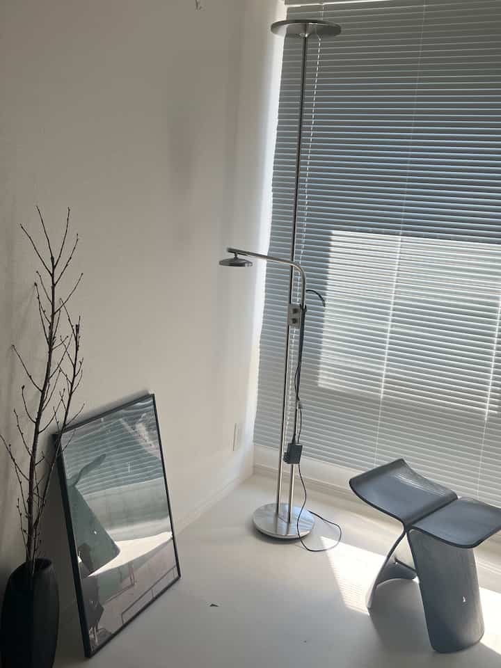 黒いスツールとシルバーのフロアランプがある狭くモダンな白い部屋、窓にブラインドが設置されたすっきりとしたインテリア