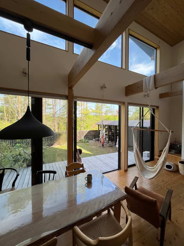 Natural brown wood and white walls define a dining room connecting to a veranda with visible children and a hanging hammock chair, creating a cozy atmosphere