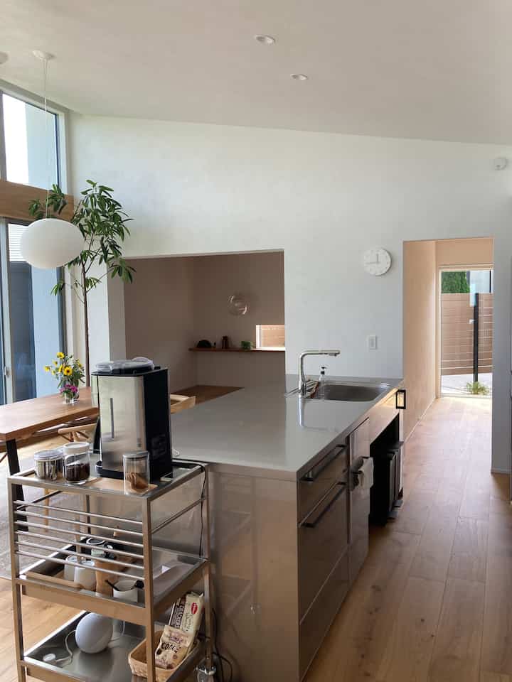 白い壁と木目調の床が調和するモダンなキッチン空間、ステンレスのアイランドキッチンとコーヒーメーカーが配置された明るく清潔感のあるインテリア