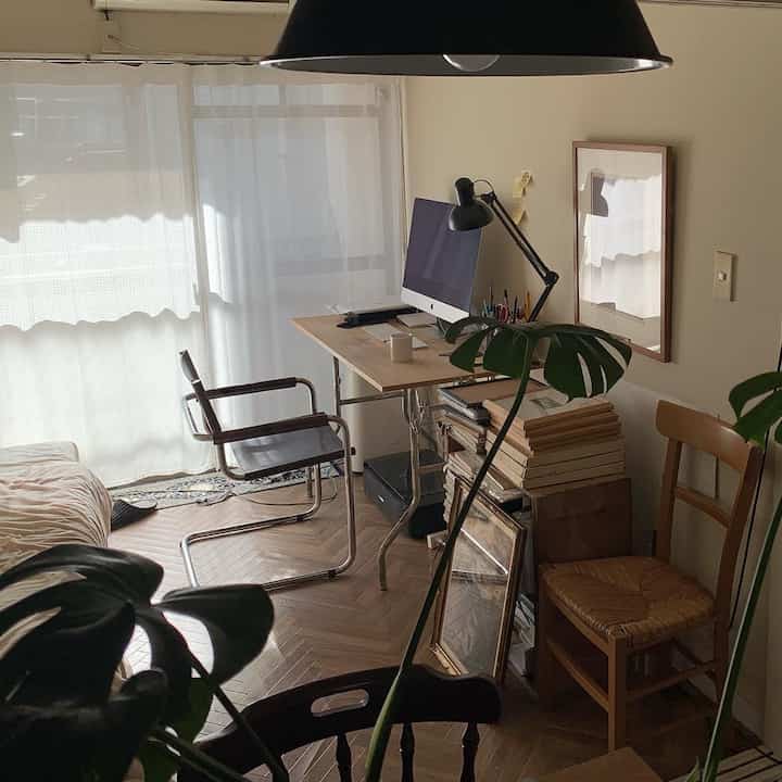 Natural toned compact home office space featuring wooden flooring, sheer curtains, and a simple desk with cozy atmosphere