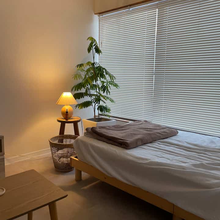 A cozy single bedroom with white blinds, wood-toned bed frame, nightstand, and table lamp arranged warmly