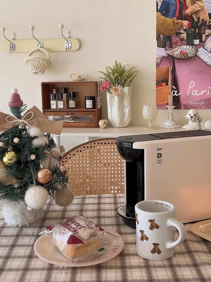 淡いベージュ色の壁とチェック柄のテーブルクロスの上にクリスマスツリー、コーヒーマシン、テディベアのマグカップが配置された雰囲気のあるホームカフェダイニング