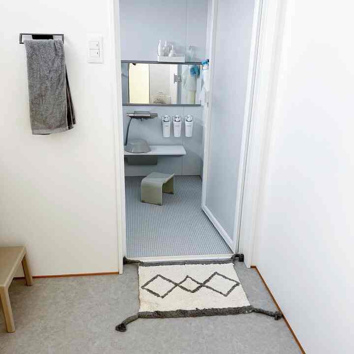 白とグレーのトーンでまとめられたバスルームの入口、ダイヤ柄のバスマットと風呂いすが配置された清潔感のある空間