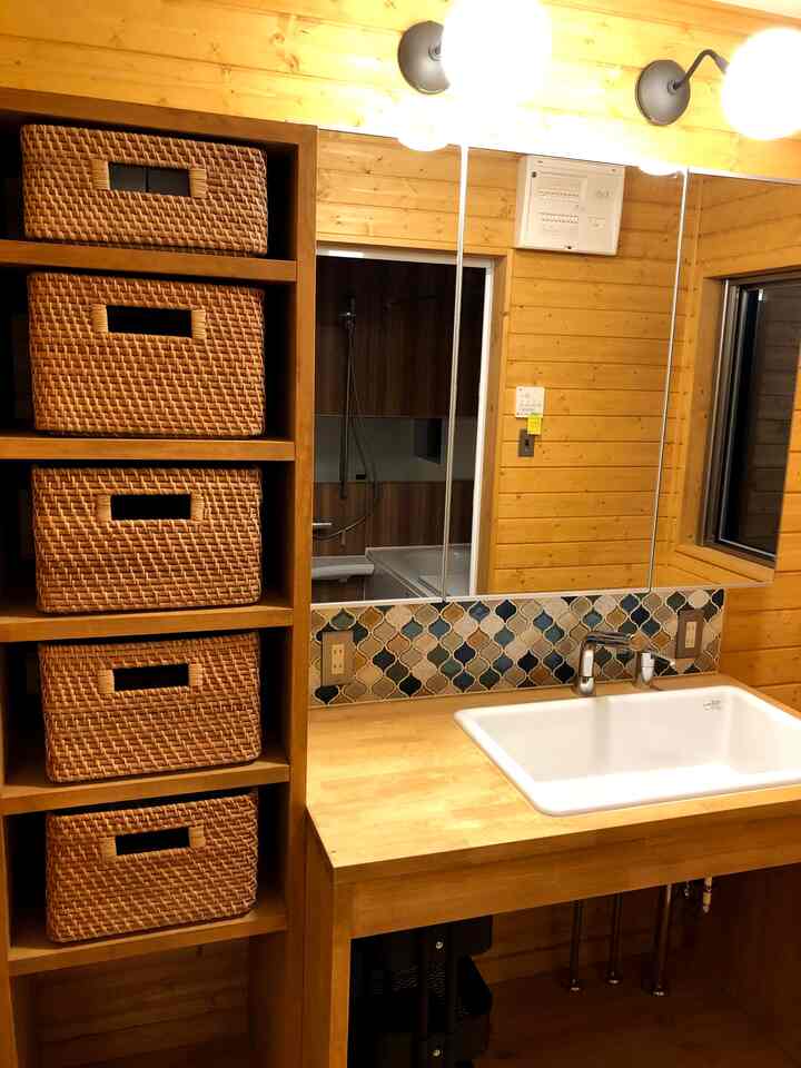 Natural wood-toned bathroom washbasin area featuring custom cabinetry, ethnic-style tile backsplash, and rattan baskets creating a neat interior