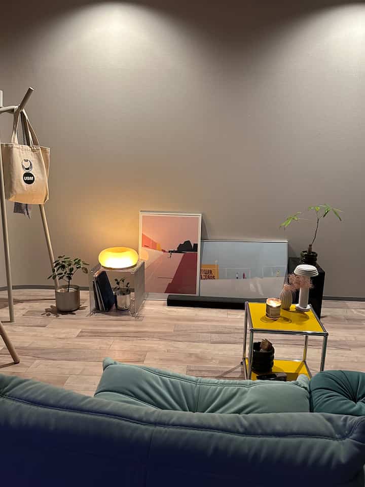 アイボリーの壁とブラウンの木製床のリビング、ミント色のソファと黄色のサイドテーブル、ポスターや照明、植物が配置された雰囲気のあるインテリア