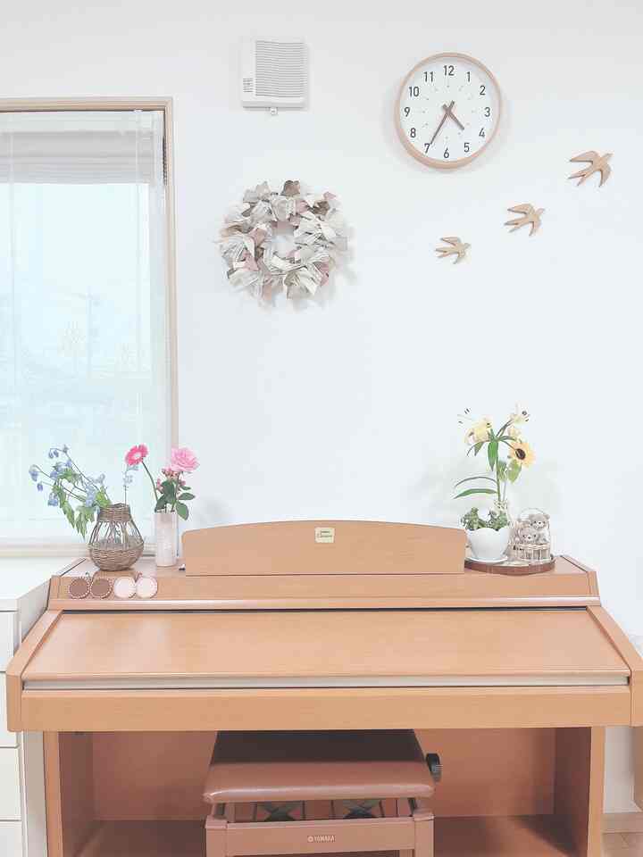 明るくナチュラルな色調のリビング、中央に木製の電子ピアノ、壁時計、花瓶と木製の鳥のオブジェが配置された雰囲気のある空間