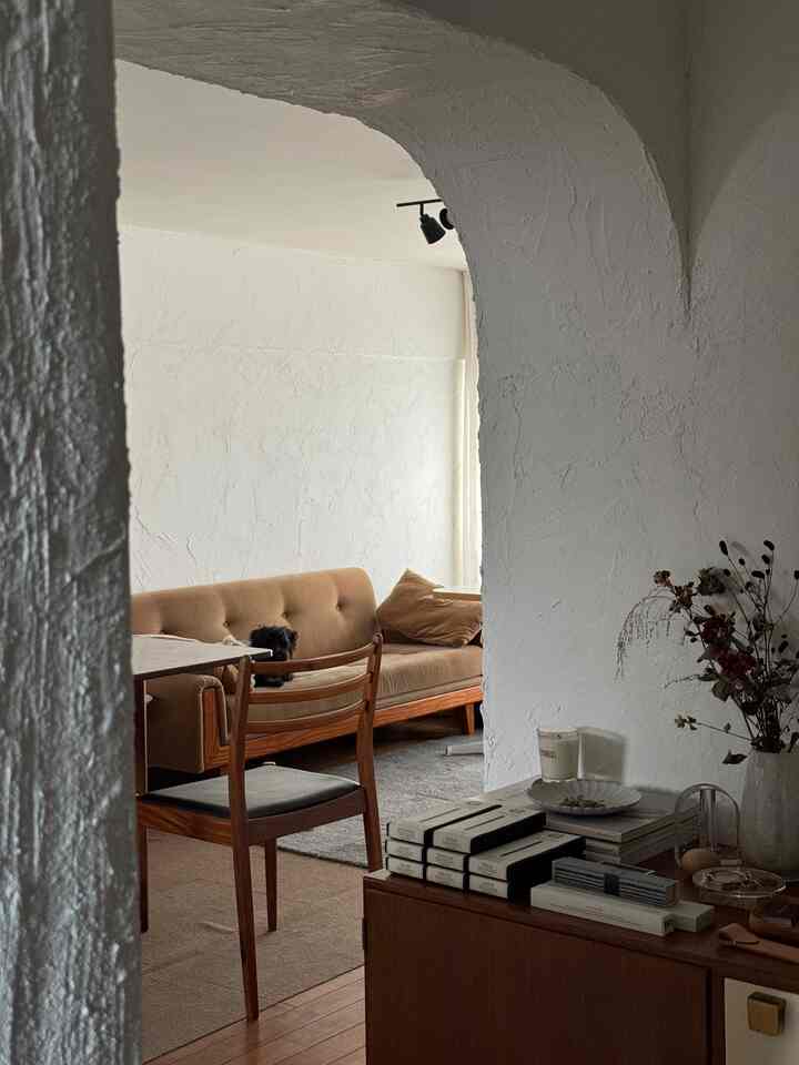 Natural-toned living room with beige sofa, wooden dining chair, white textured walls, and arched doorway