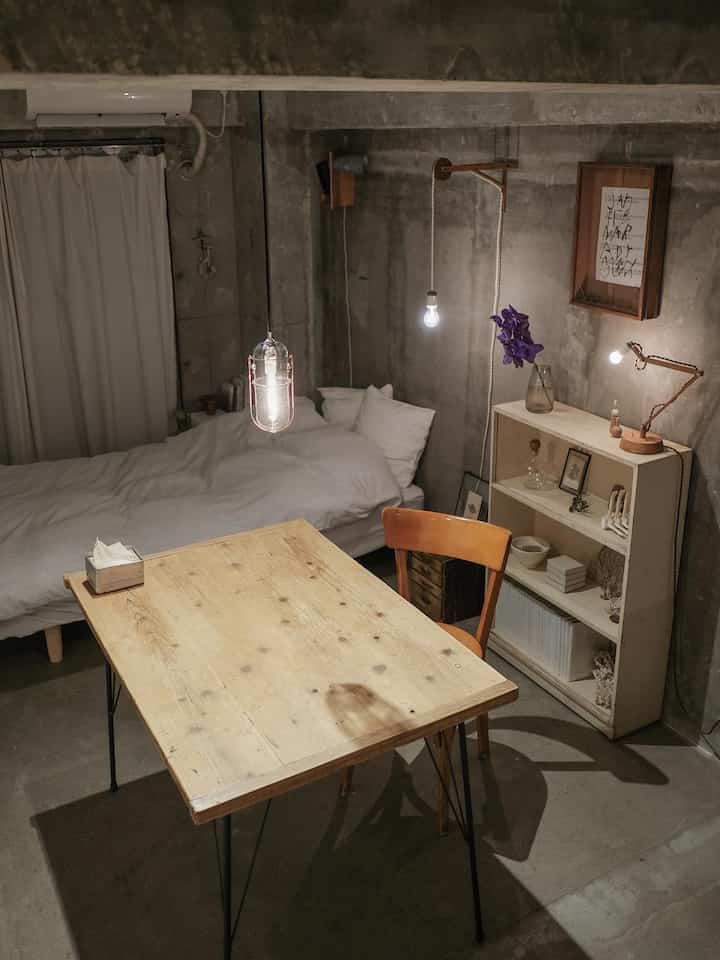 グレーのコンクリート壁とナチュラルな木製家具が配置されたワンルーム寝室、ペンダントライトとテーブルランプが温かみのある雰囲気を演出