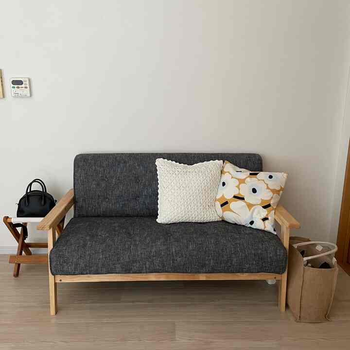 Natural-toned two-seater sofa with cushions, small stool on left and eco bag on right in a simple living room space