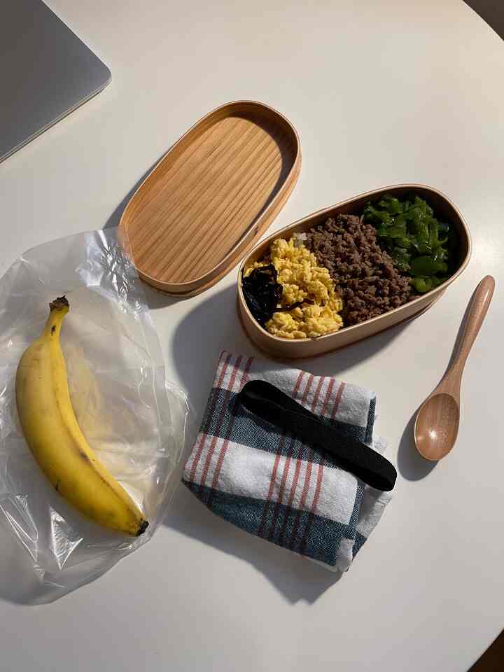 白いテーブルの上に置かれた木目調の弁当箱とバナナ、木製スプーンとチェック柄の布が配置された清潔感のある食事空間